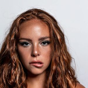 Studio headshot of a young woman with curly red hair on a white background.